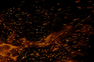 Bright orange and yellow sparks fly against a dark black background, creating a dynamic and fiery effect that resembles embers or fire particles in motion.