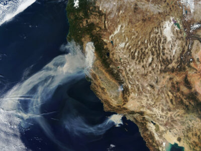 A satellite image showing smoke plumes from wildfires spreading across parts of the western United States, mainly affecting California. The smoke is drifting over the Pacific Ocean, while the land shows varying terrain and weather patterns.