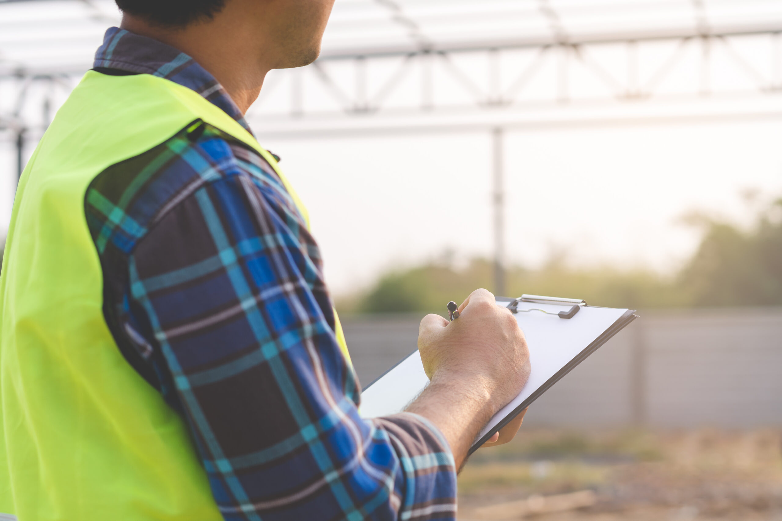 A person in a safety vest holds a clipboard and appears to be writing. They are standing at a construction site with a steel framework structure in the background, and the scene is lit by natural daylight.