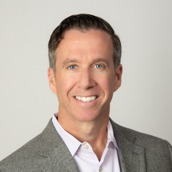 A smiling man with short dark hair is wearing a light gray suit jacket over a white dress shirt. He is posed against a neutral background, looking directly at the camera.