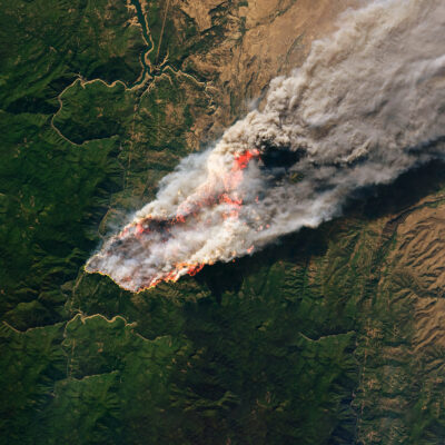 Aerial view of a large wildfire engulfing a forested region, with thick smoke billowing into the sky. The flames are spreading across green terrain, leaving scorched earth in their path.