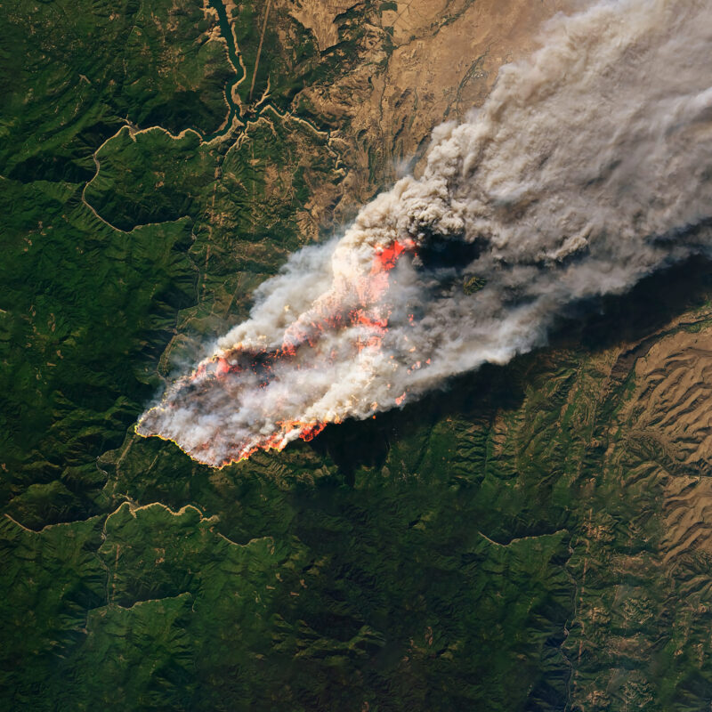 Aerial view of a large wildfire engulfing a forested region, with thick smoke billowing into the sky. The flames are spreading across green terrain, leaving scorched earth in their path.