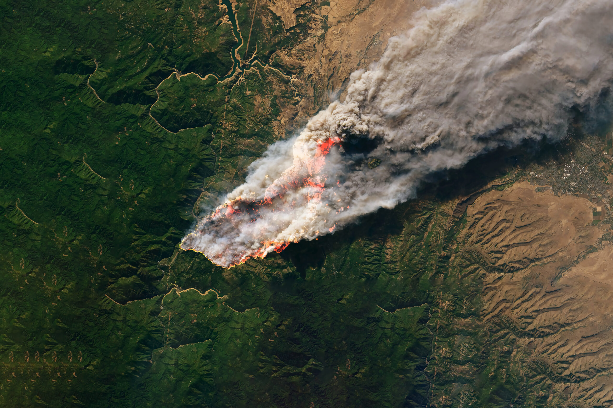 Aerial view of a large wildfire engulfing a forested region, with thick smoke billowing into the sky. The flames are spreading across green terrain, leaving scorched earth in their path.