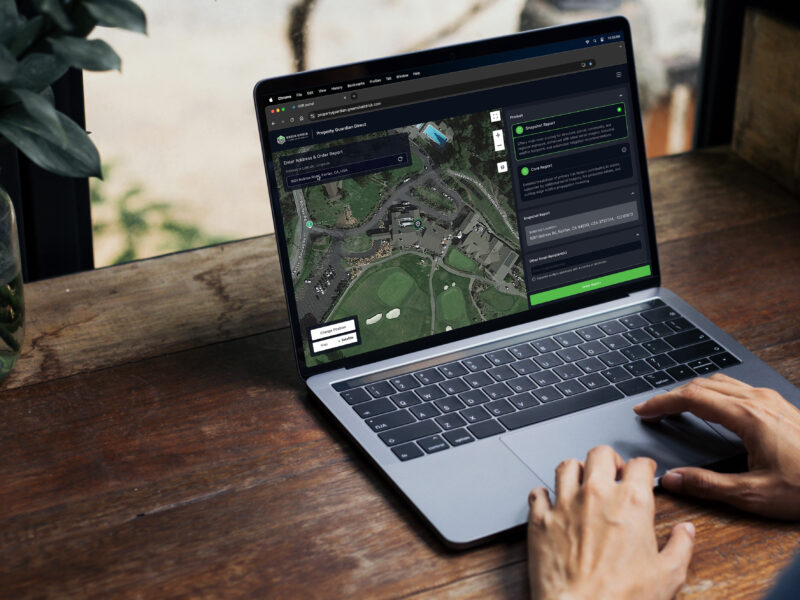 Person using a laptop displaying a map with a chat interface. The screen shows a satellite view of a landscape. The person is seated at a wooden table near a window with plants in the background.