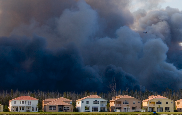 A row of suburban houses stands in front of a forest fire, with thick, dark smoke billowing into the sky. A small aircraft is flying above the smoke, likely monitoring or responding to the fire.