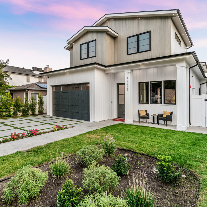 Modern two-story house with gray garage door, white exterior, and black trim. Front yard features a manicured lawn, flower beds, and a small patio with two chairs and a table. The sky is colorful at sunset.