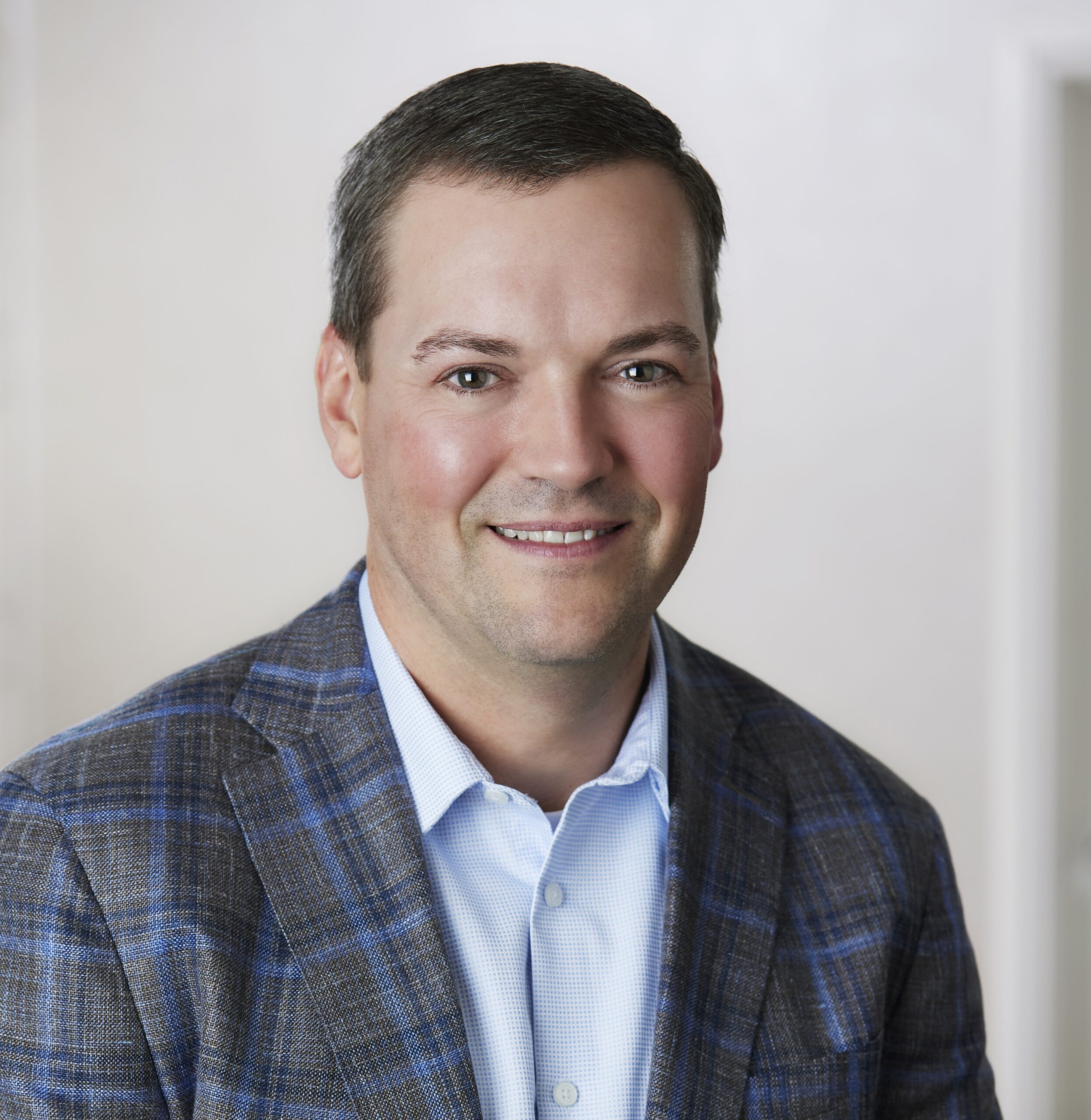 A man with short dark hair, wearing a blue plaid suit jacket and a light blue collared shirt, smiles at the camera against a neutral indoor background.