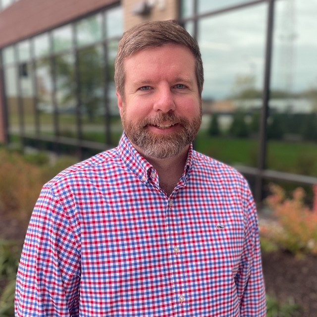 A man with short brown hair and a beard, wearing a red, white, and blue checkered shirt, stands smiling outside in front of a building with large windows and landscaping.