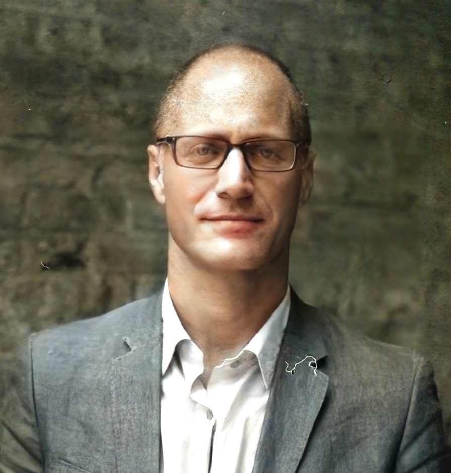 A middle-aged man with short hair and glasses, wearing a gray suit jacket and white shirt, stands in front of a textured, dark background, looking at the camera with a neutral expression.