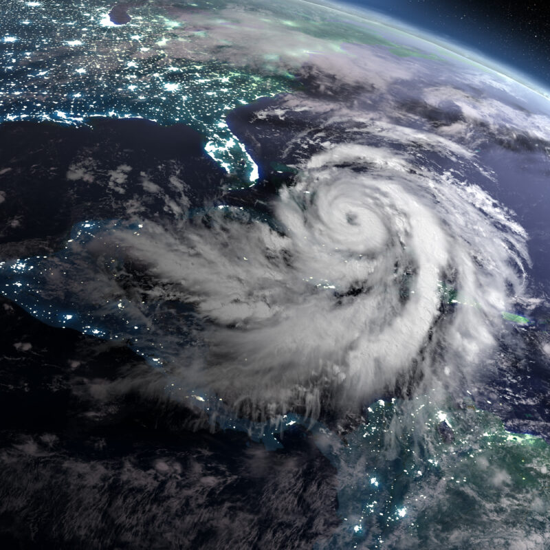 A large hurricane with spiral-shaped clouds is seen over the Gulf of Mexico from space, approaching the southeastern United States. City lights are visible along the coastline beneath the storm.
