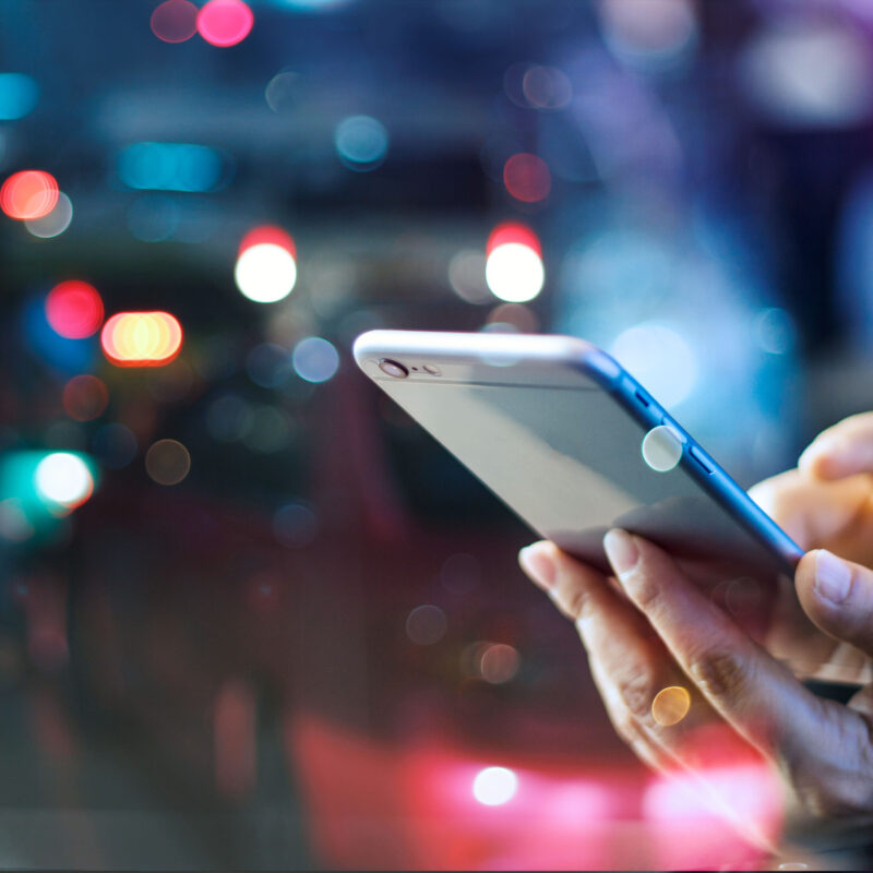 Close-up of a person’s hands using a smartphone at night, with colorful city lights blurred in the background.