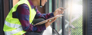 A person wearing a safety vest and helmet crouches next to a metal fence, holding a clipboard and pen, appearing to inspect or take notes.