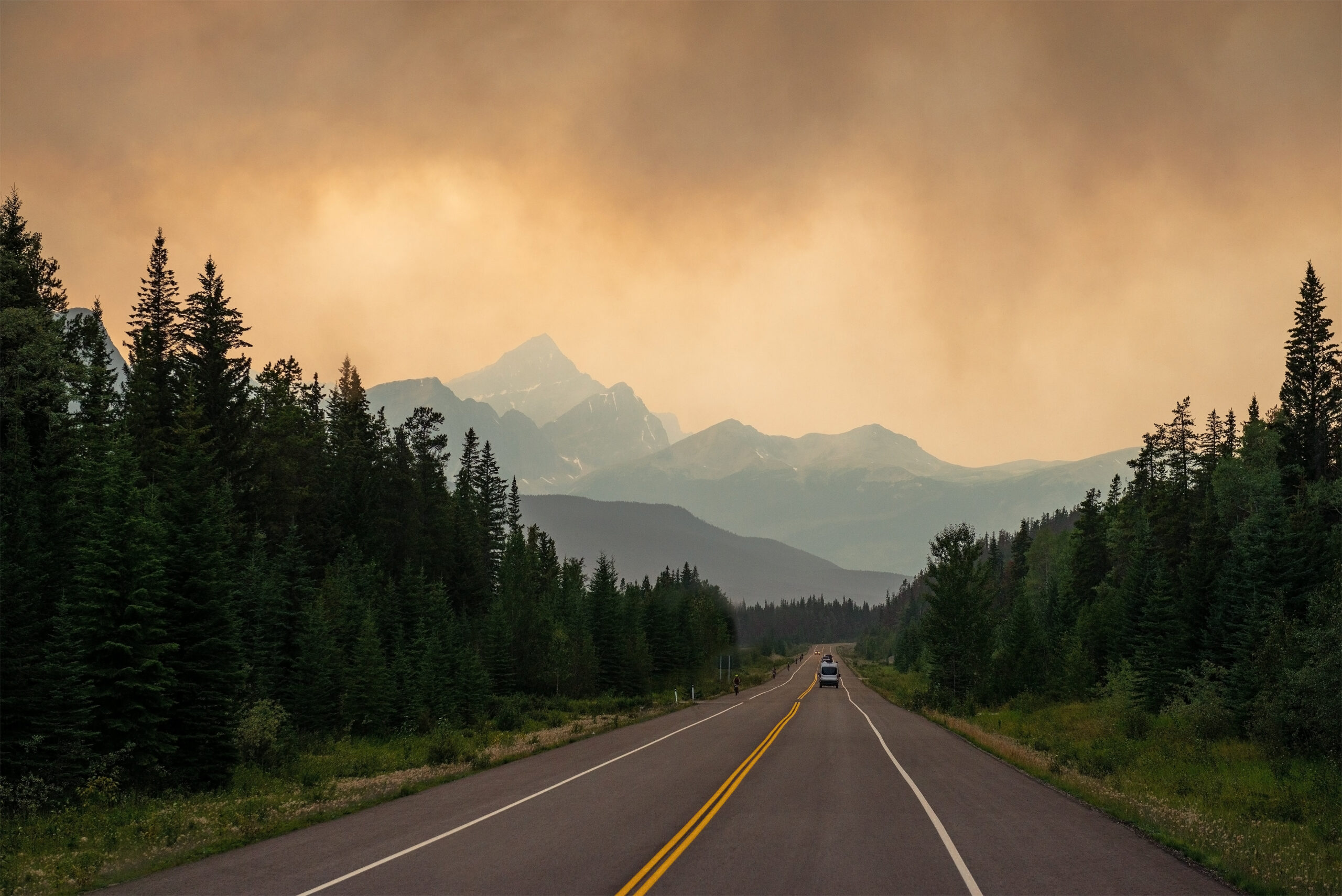 A two-lane road runs through a forest with tall trees on both sides, leading towards distant mountains under a smoky orange sky, with a few cars driving ahead.