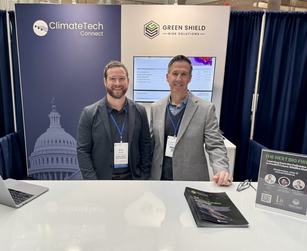 Two men in business attire stand smiling at a trade show booth for ClimateTech Connect and Green Shield Risk Solutions. There are banners, brochures, a laptop, and a display screen behind them.