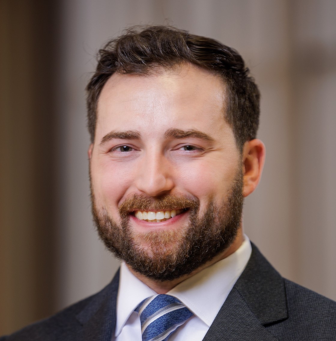 A man with short dark hair and a full beard, wearing a dark suit, white shirt, and striped tie, smiling at the camera against a blurred indoor background.