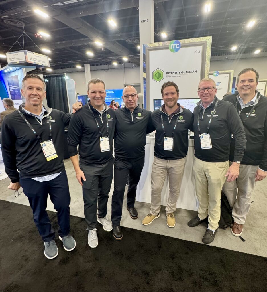 Six men stand together at a convention booth labeled "Property Guardian" at ITC. They are smiling, wearing matching dark jackets, and have name badges, with an exhibition hall in the background.