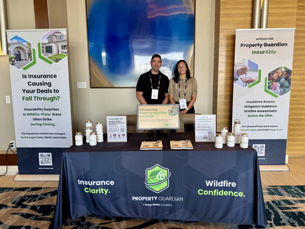 Two people stand behind a promotional table for Property Guardian insurance. The table displays brochures and jars. Banners on both sides highlight insurance clarity, wildfire confidence, and insurability in wildfire-prone areas.