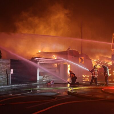 Firefighters spray water on a burning building at night, with heavy smoke and flames visible. Emergency lights shine, and hoses are spread across the wet street as crews work to control the blaze.