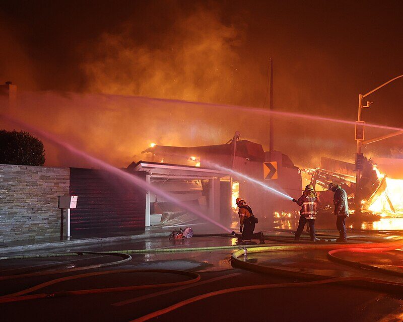 Firefighters spray water on a burning building at night, with heavy smoke and flames visible. Emergency lights shine, and hoses are spread across the wet street as crews work to control the blaze.