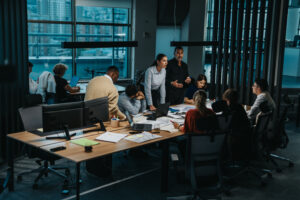 A group of people are gathered around a table in a modern office, engaged in a discussion with laptops and documents. Large windows show a cityscape outside. The atmosphere appears focused and collaborative.