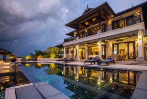 A luxurious two-story villa with large windows and balconies, warmly lit at dusk, sits beside a serene swimming pool surrounded by lounge chairs under a dramatic, cloudy sky.