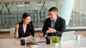Two business professionals sit at a desk discussing documents and charts. A laptop, notebook, coffee mugs, and a small plant are on the table. Both are smiling and engaged in conversation in a modern office setting.