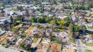 Aerial view of a residential neighborhood showing widespread destruction from a wildfire; most houses are reduced to rubble and foundations, with some trees and a few undamaged buildings visible in the background.