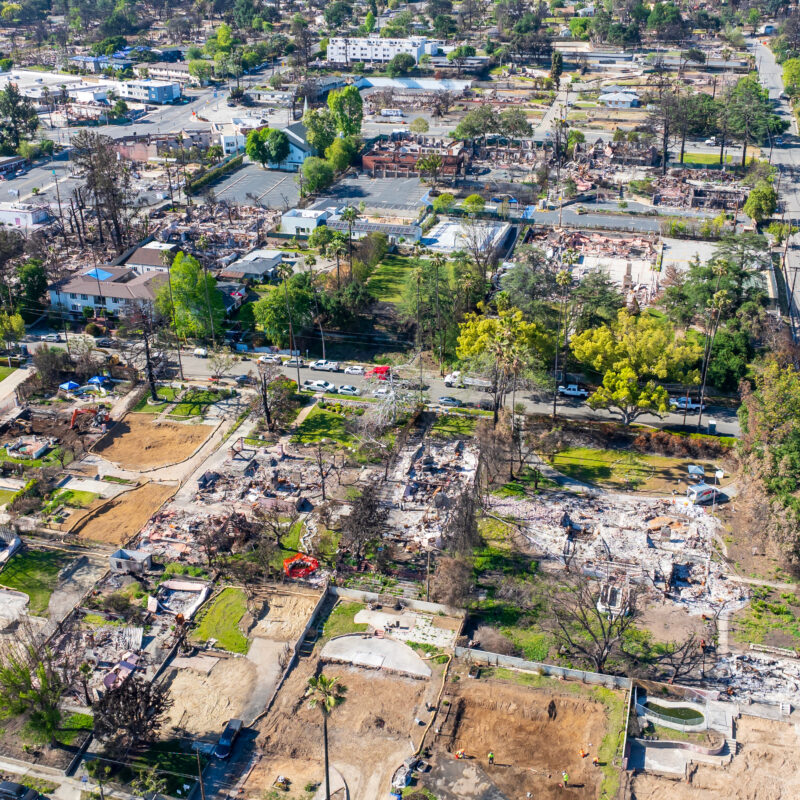 Aerial view of a residential neighborhood showing widespread destruction from a wildfire; most houses are reduced to rubble and foundations, with some trees and a few undamaged buildings visible in the background.