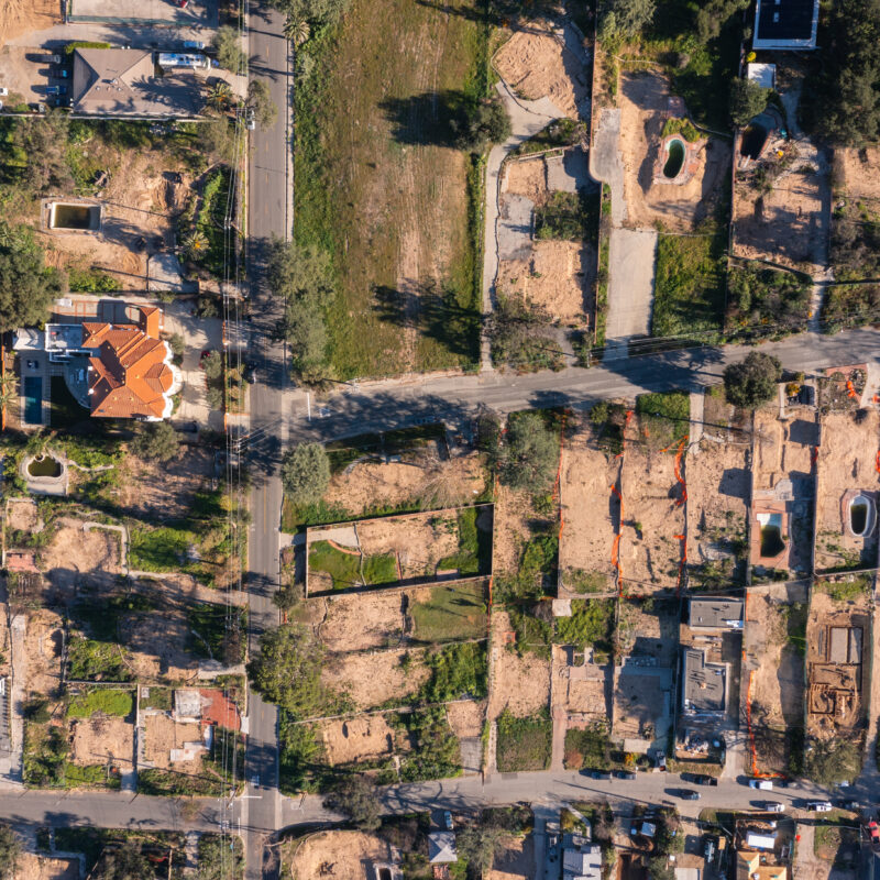 Aerial view of a suburban neighborhood with houses, roads, and many empty plots of land or construction sites, seen from above in daylight. Trees and cars are visible around the area.