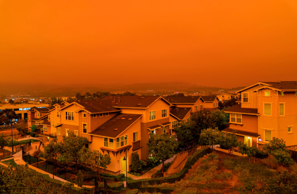 Rows of suburban houses are shown under an intense orange sky, likely due to wildfire smoke, creating an eerie and dramatic atmosphere over the residential neighborhood.