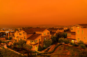 Rows of suburban houses are shown under an intense orange sky, likely due to wildfire smoke, creating an eerie and dramatic atmosphere over the residential neighborhood.