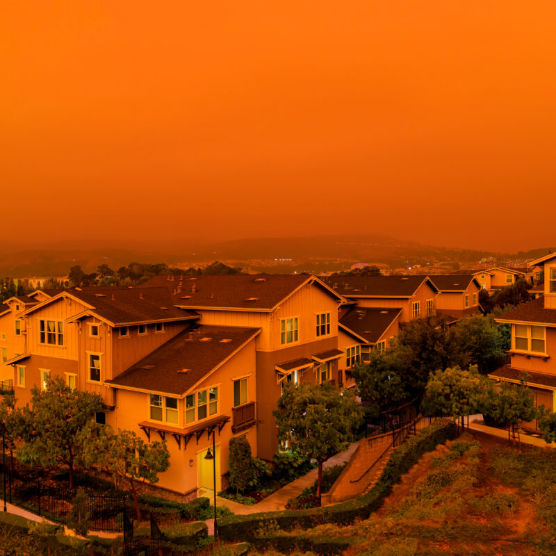 Rows of suburban houses are shown under an intense orange sky, likely due to wildfire smoke, creating an eerie and dramatic atmosphere over the residential neighborhood.