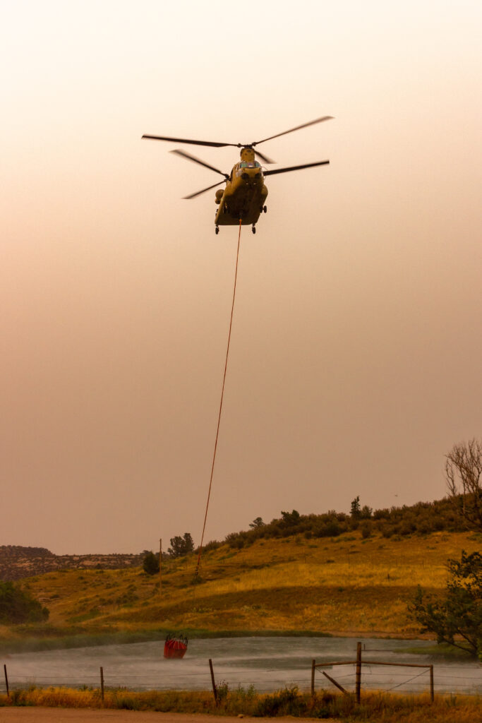 A helicopter flies above a grassy field at sunset, carrying a large red water bucket suspended by a long cable, likely for firefighting purposes.