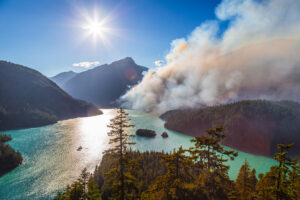 A bright sun shines over a turquoise lake surrounded by forested mountains, while thick smoke rises from a wildfire in the trees along the shoreline under a clear blue sky.