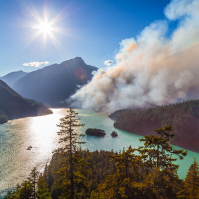 A bright sun shines over a turquoise lake surrounded by forested mountains, while thick smoke rises from a wildfire in the trees along the shoreline under a clear blue sky.