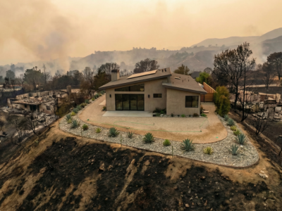 A modern house sits untouched on a hilltop surrounded by scorched earth and burnt remains of other homes, with smoke rising in the background from a recent wildfire.