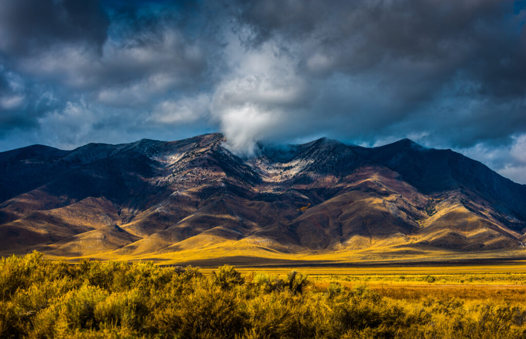 Dramatic clouds hover over a rugged mountain range with sunlight illuminating the golden-yellow foothills and grassy plains in the foreground.