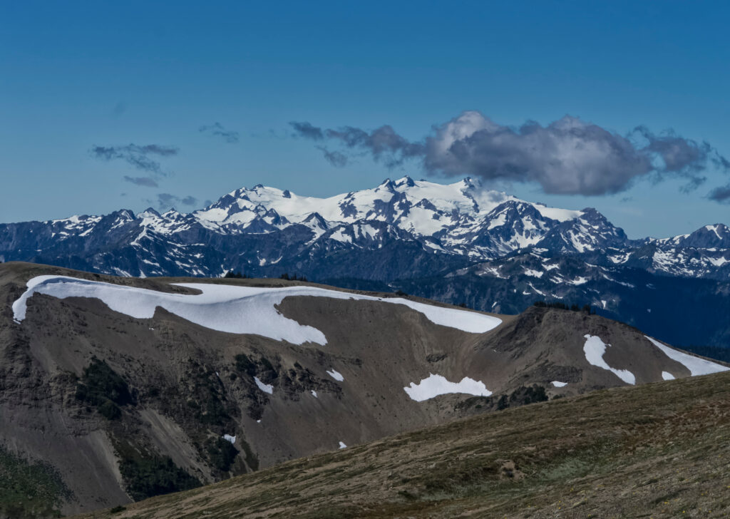 Snow-capped mountain range under a clear blue sky, with patches of snow on rocky and grassy foreground hills untouched by wildfire. A single fluffy cloud hovers above the distant peaks.