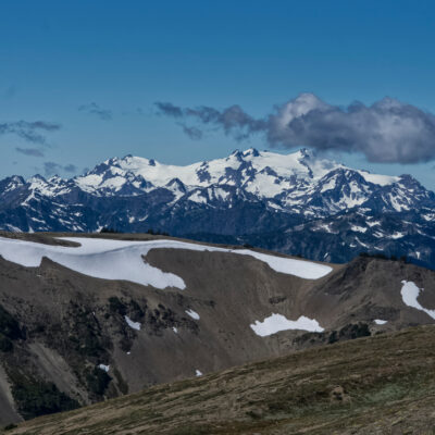 Snow-capped mountain range under a clear blue sky, with patches of snow on rocky and grassy foreground hills untouched by wildfire. A single fluffy cloud hovers above the distant peaks.