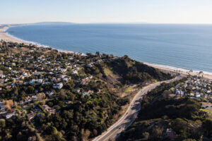 Aerial view of a coastal area with winding roads, green hills touched by a recent wildfire, residential homes, and a sandy beach bordering the blue ocean under a clear sky.