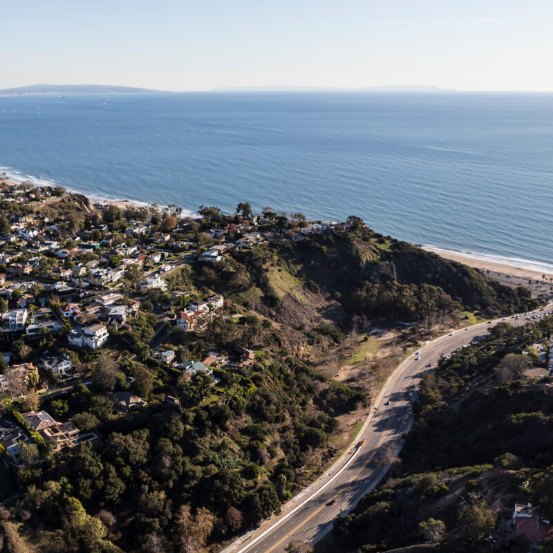 Aerial view of a coastal area with winding roads, green hills touched by a recent wildfire, residential homes, and a sandy beach bordering the blue ocean under a clear sky.