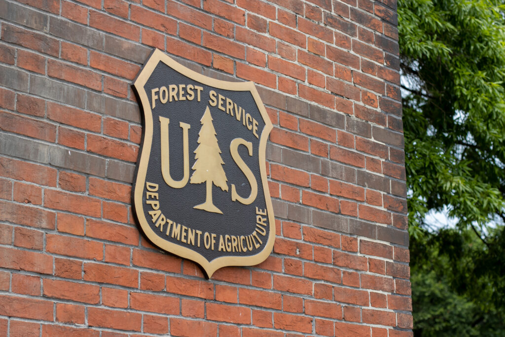 A bronze and black U.S. Forest Service Department of Agriculture shield is mounted on a red brick wall, with green tree branches visible to the right—a reminder of the agency’s vital role in wildfire prevention.