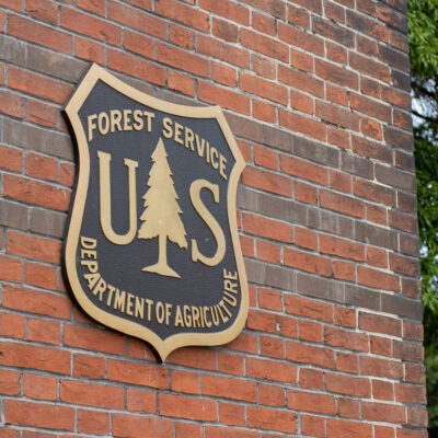 A bronze and black U.S. Forest Service Department of Agriculture shield is mounted on a red brick wall, with green tree branches visible to the right—a reminder of the agency’s vital role in wildfire prevention.