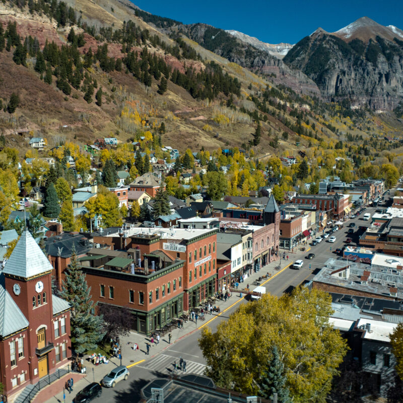 Aerial view of a small mountain town with historic brick buildings, tree-lined streets, and colorful autumn foliage, surrounded by forested hills recovering from past wildfire and snow-capped peaks under a clear blue sky.