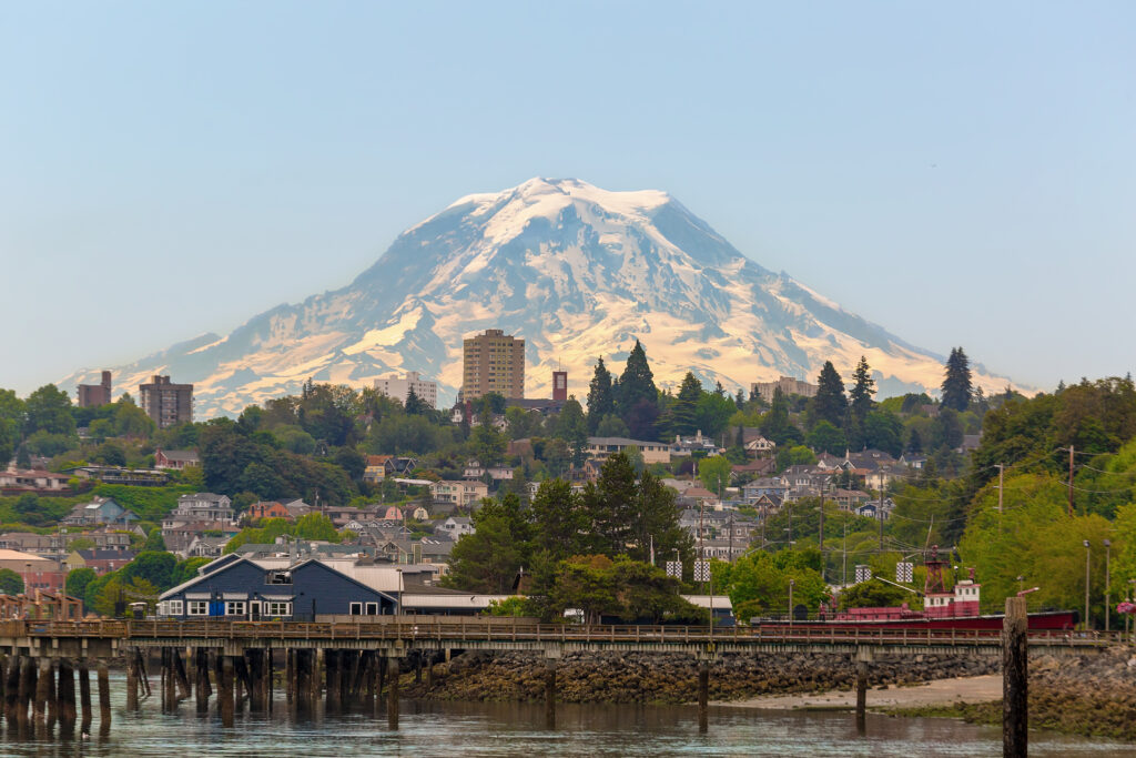 A scenic view of a waterfront city with a wooden pier, houses, and tall buildings, set against a backdrop of a large snow-capped mountain under a clear sky recovering from recent wildfire.