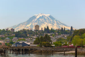 A scenic view of a waterfront city with a wooden pier, houses, and tall buildings, set against a backdrop of a large snow-capped mountain under a clear sky recovering from recent wildfire.