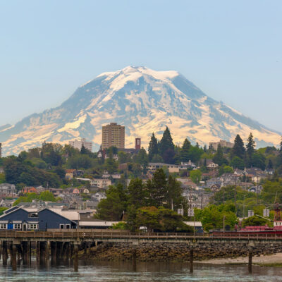 A scenic view of a waterfront city with a wooden pier, houses, and tall buildings, set against a backdrop of a large snow-capped mountain under a clear sky recovering from recent wildfire.