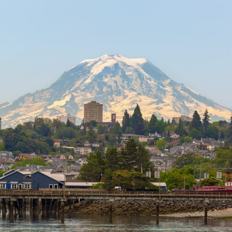 A scenic view of a waterfront city with a wooden pier, houses, and tall buildings, set against a backdrop of a large snow-capped mountain under a clear sky recovering from recent wildfire.
