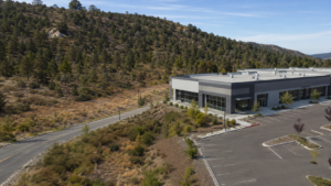 A modern gray commercial building sits next to a mostly empty parking lot, bordered by a winding road and a hillside covered in trees and dry grass—landscape vulnerable to wildfire—under a clear blue sky.