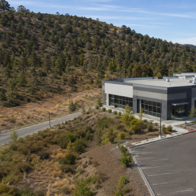 A modern gray commercial building sits next to a mostly empty parking lot, bordered by a winding road and a hillside covered in trees and dry grass—landscape vulnerable to wildfire—under a clear blue sky.